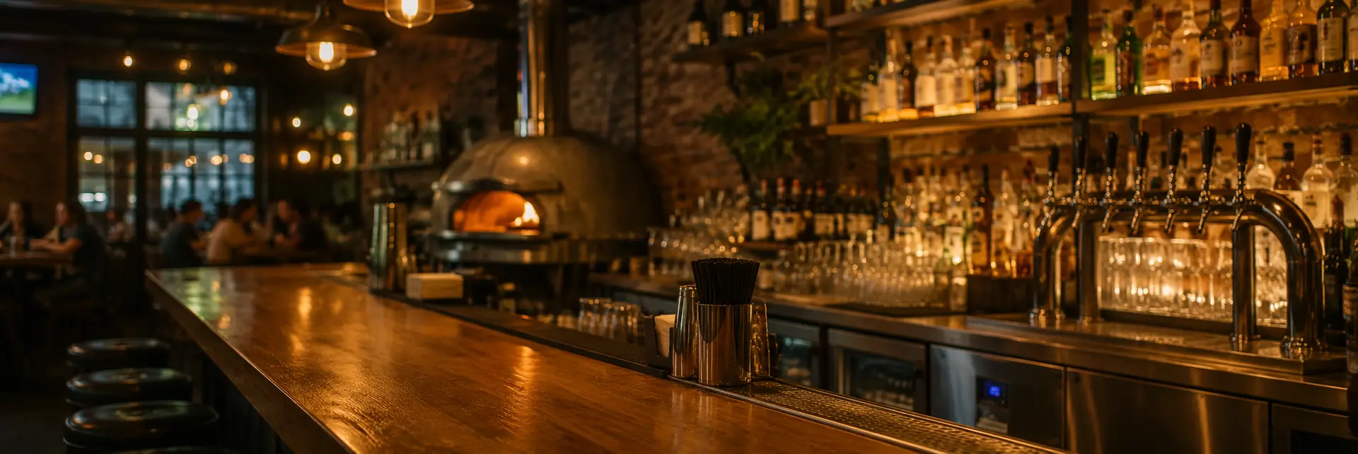 Balcão de bar em madeira com torres de chope, prateleira de garrafas iluminada e forno de pizza ao fundo, com clientes nas mesas do salão